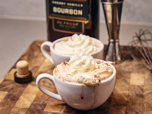 Bourbon hot cocoa topped with whipped cream served in white mugs beside St. Elmo Cherry Vanilla Bourbon.