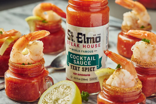 Small shrimp cocktail shooters filled with spicy cocktail sauce and topped with shrimp, served around a bottle of St. Elmo Steak House Cocktail Sauce with fresh lime wedges on a marble surface.