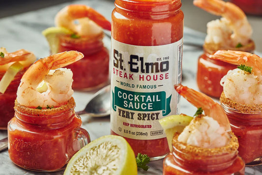 Small shrimp cocktail shooters filled with spicy cocktail sauce and topped with shrimp, served around a bottle of St. Elmo Steak House Cocktail Sauce with fresh lime wedges on a marble surface.