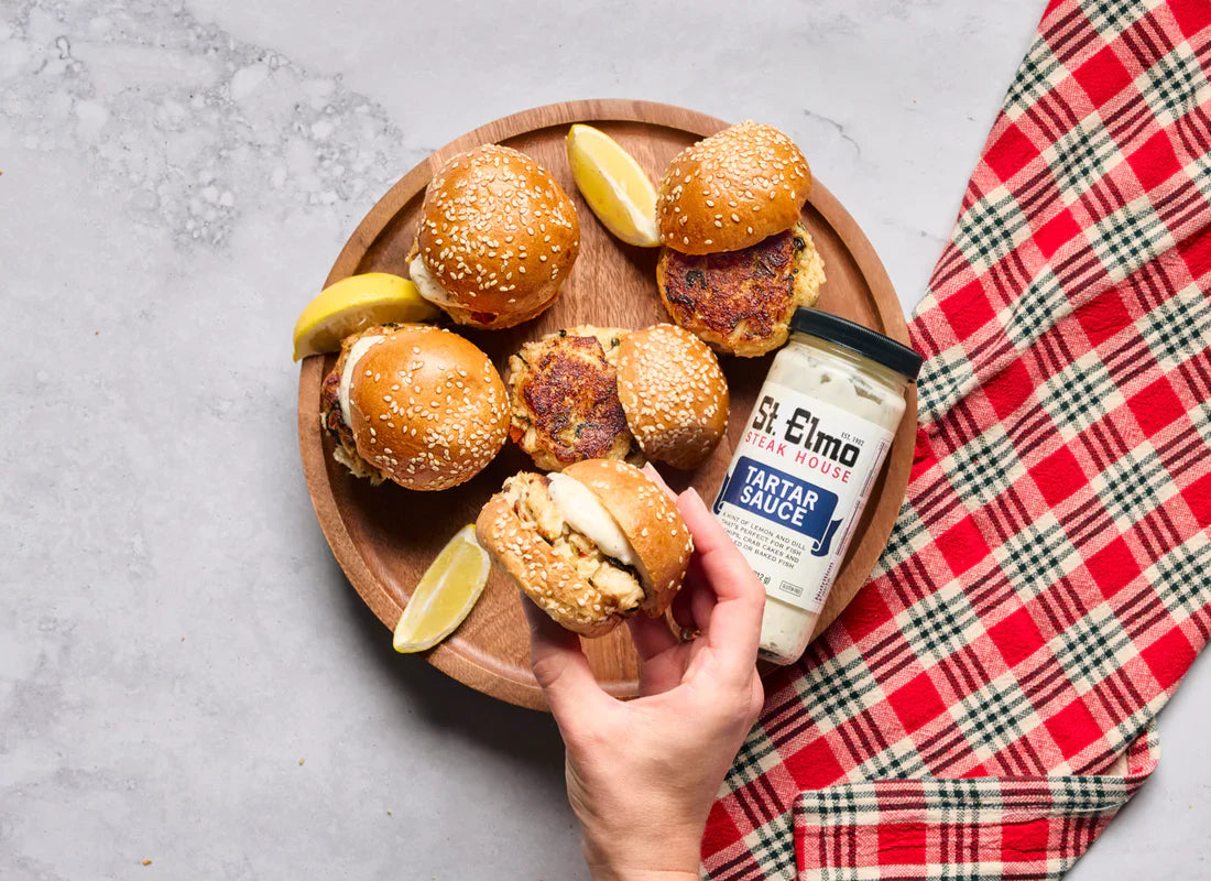 Crab cake sliders on sesame seed buns served on a wooden platter with lemon wedges and a jar of St. Elmo Steak House Tartar Sauce, with a hand holding one slider above the plate.