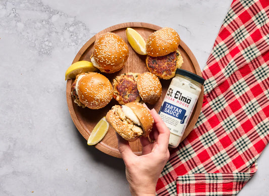 Crab cake sliders on sesame seed buns served on a wooden platter with lemon wedges and a jar of St. Elmo Steak House Tartar Sauce, with a hand holding one slider above the plate.
