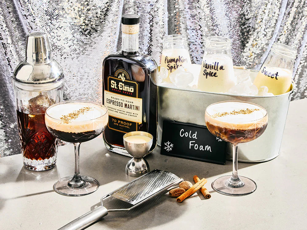 Two eggnog espresso martinis topped with creamy cold foam and cinnamon garnish beside a bottle of St. Elmo Espresso Martini and holiday cocktail ingredients.
