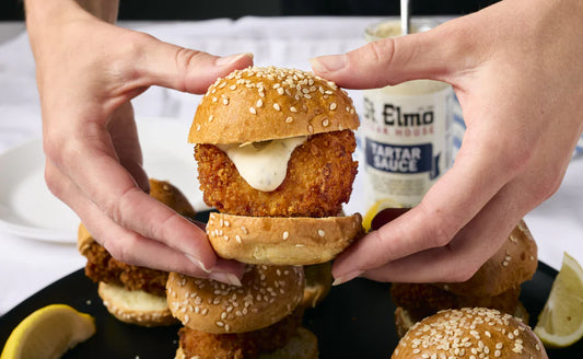 Crispy fried shrimp slider on a sesame seed bun topped with tartar sauce, surrounded by mini shrimp sandwiches and lemon wedges.