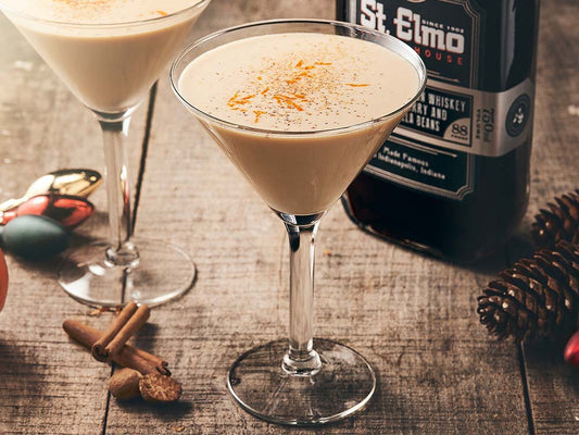 Creamy bourbon eggnog martini garnished with freshly grated nutmeg, cinnamon, and orange zest in a martini glass beside a bottle of cherry vanilla bourbon.