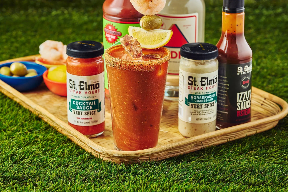 Spicy Bloody Mary garnished with shrimp, lemon, and blue cheese olives, served with cocktail sauce, horseradish, and hot sauce on a wooden tray.
