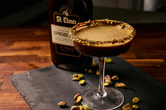 Espresso martini with a chopped pistachio rim served in a coupe glass beside a bottle of espresso martini cocktail and scattered pistachios.