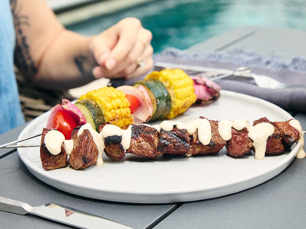 Grilled steak kebabs drizzled with creamy sauce and served with vegetable skewers featuring corn, zucchini, onion, and tomatoes.