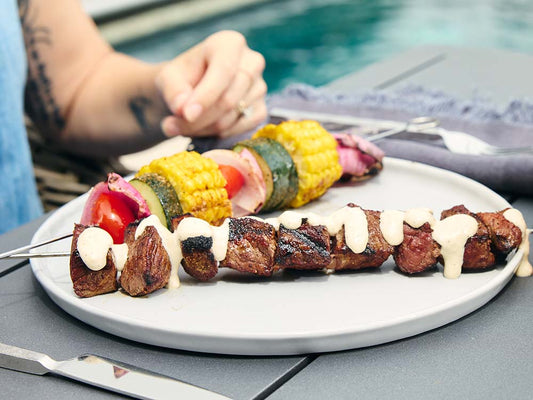 Grilled steak kebabs drizzled with creamy sauce and served with vegetable skewers featuring corn, zucchini, onion, and tomatoes.