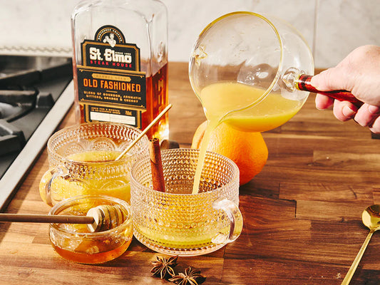 Warm old fashioned hot toddy being poured into a glass mug, garnished with cinnamon sticks, star anise, and orange, beside a bottle of barrel-aged old fashioned cocktail.