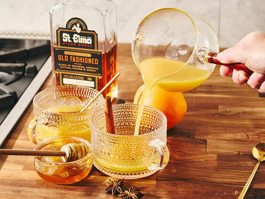 Warm old fashioned hot toddy being poured into a glass mug, garnished with cinnamon sticks, star anise, and orange, beside a bottle of barrel-aged old fashioned cocktail.