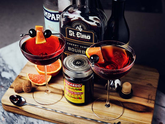 Sparkling Boulevardier cocktail garnished with orange wedges and cherries, served in coupe glasses beside cherry vanilla bourbon, Campari, and vermouth.