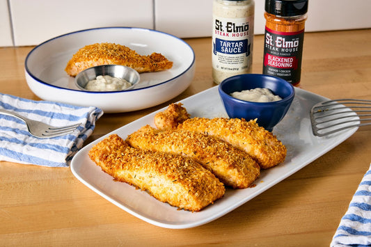Parmesan-crusted salmon fillets coated with crispy breadcrumbs and blackened seasoning, served with tartar sauce alongside St. Elmo seasoning bottles on a wooden table.