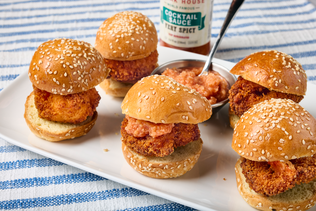 Crispy shrimp sliders on sesame seed buns topped with St. Elmo Cocktail Sauce and served on a white platter.