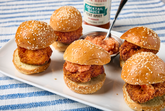 Crispy shrimp sliders on sesame seed buns topped with St. Elmo Cocktail Sauce and served on a white platter.